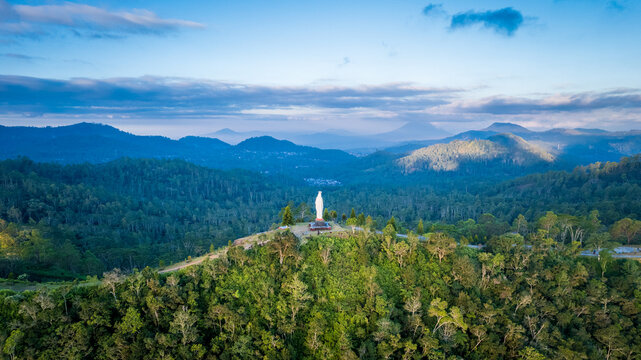 Aerial view of the Virgin Mary statue standing atop a lush green hill surrounded by dense forest and rolling mountains under a clear blue sky in Bajawa, Nusa Tenggara Timur, Indonesia.