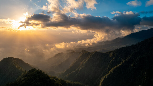 Aerial view of lush green mountains covered in dense forest under a dramatic sunset sky with golden light filtering through clouds in Bajawa, Nusa Tenggara Timur, Indonesia.
