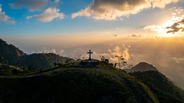 Aerial view of a Christian cross standing on a hilltop at sunset with golden light illuminating the surrounding mountains and clouds in Bajawa, Nusa Tenggara Timur, Indonesia.