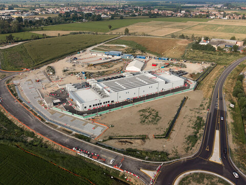 Aerial view of the BYES MIL08 data center construction site with a large white building, surrounding dirt areas, construction equipment, and a nearby road in Siziano, Lombardia, Italy.