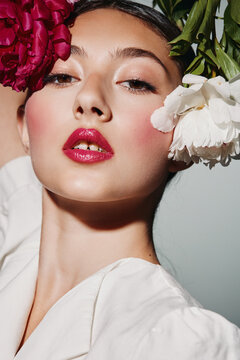 Close-up of fashion model with vibrant makeup and glossy red lips surrounded by large pink and white flowers. Creative beauty portrait with artistic floral styling on white background.