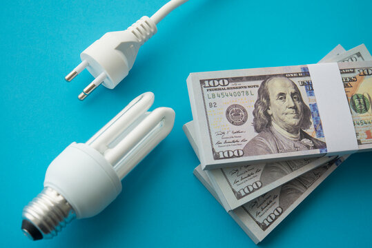 Electrical plug, light bulb, heap US banknotes on blue background. Electricity bill prices increase, energy crisis, home or business financial management and planning, expense budget control concept.