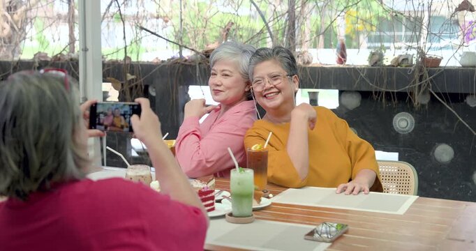 Senior Asian women friends pose for smartphone photo at cafe table during joyful reunion, smiling after long time apart while sharing dessert, sweet drink, companionship, encouragement, warm memory.