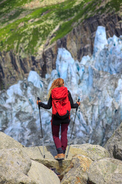 Rear view of female hiker with red backpack looking at massive glacier in mountains- Alps