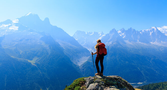 Rear view of female hiker with red backpack standing on rock looking at majestic alpine mountain range and mont blanc