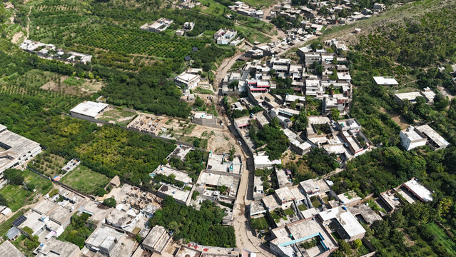 Aerial view of Dir District with dense housing, narrow dirt roads, and surrounding lush green orchards in the mountainous terrain of Khyber Pakhtunkhwa, Pakistan.