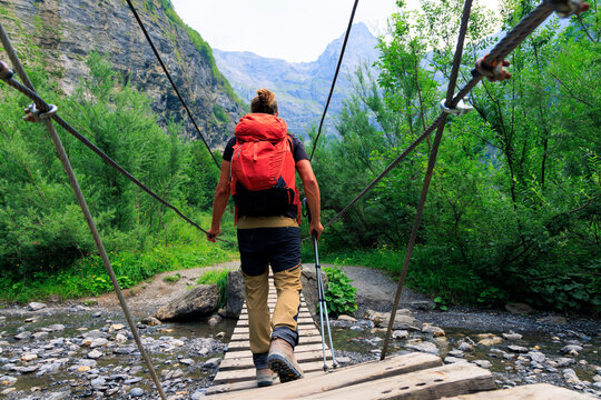 Rear view of male hiker with backpack walking on suspension footbridge over river in mountain canyon