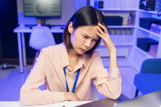 Stressed young Asian office worker feeling overwhelmed with paperwork and workload while working on laptop in office.