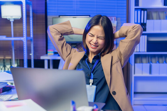 Tired young Asian businesswoman stretching at desk while working on laptop in office.