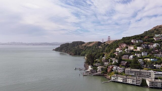 Scenic aerial view flying along the Sausalito coastline and hillside homes, with the iconic Golden Gate Bridge and San Francisco skyline visible in the background across the bay