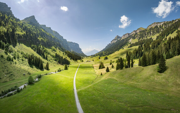 Aerial view of the Justistal valley featuring lush green meadows, steep rocky mountain slopes, dense pine forests, and a narrow road winding through the landscape in Sigriswil, Bern, Switzerland.