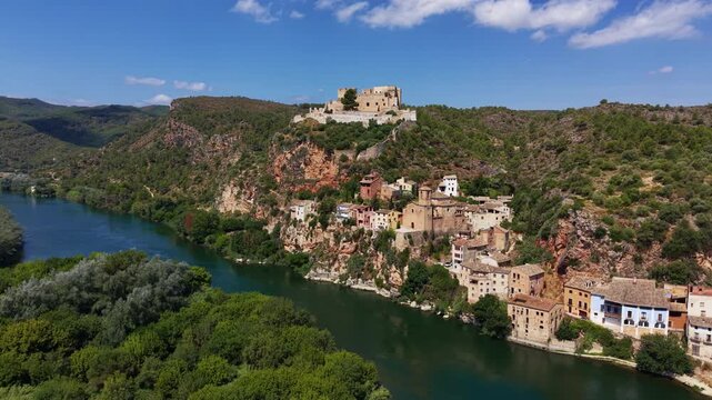 Miravet village and medieval castle perched on rocky cliffs above the Ebro River, Catalonia, Spain, surrounded by green hills and blue sky