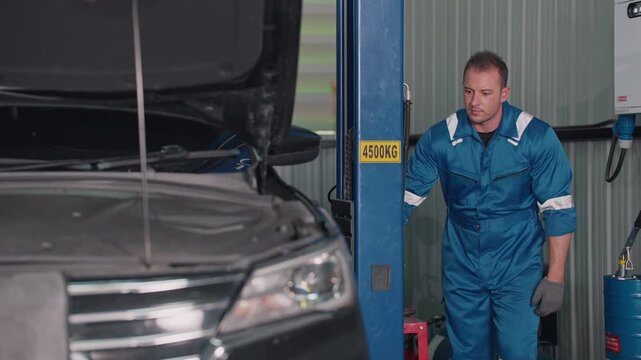 Mechanic in blue overalls works under EV car lift. He handles equipment near silver SUV's side. Focused on task, no visible emotion. Ideal for auto repair, maintenance, or service branding