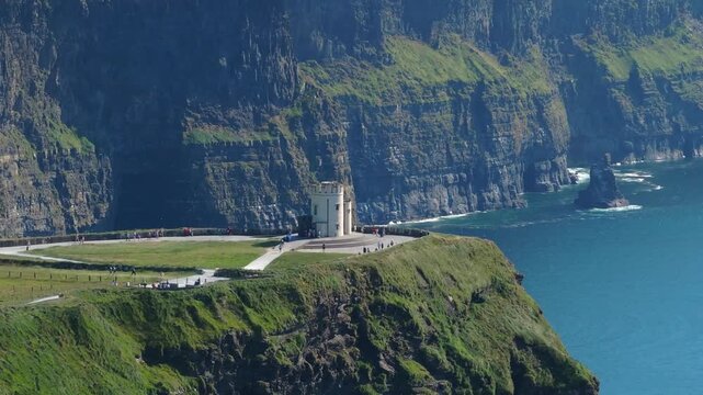 Tourists walk around OBrien Tower on a grassy headland as sheer ocean cliffs stretch along the Moher coastline in bright daylight.