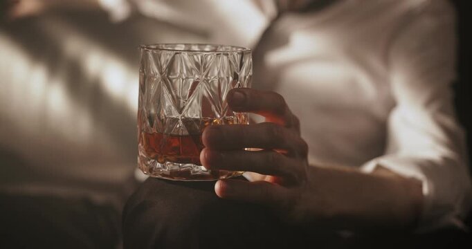 Close-up of a man holding a glass of strong alcoholic drink while sitting alone in a dark room.