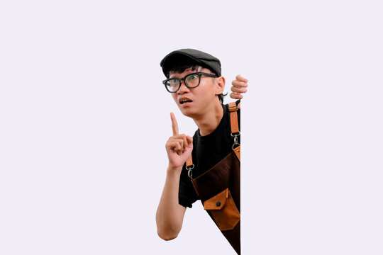 Barista Pria muda memakai kacamata dan topi, menunjuk ke atas. Dia memberi sinyal perhatian dengan ekspresi serius dan fokus. Celemek kulit coklat menunjukkan pekerjaan langsung dan praktis