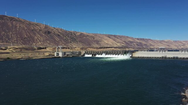 4K aerial drone footage of John Day Dam near Rufus, Oregon, USA with distant wind turbines. Hydroelectric power station, renewable energy, pelicans fishing in Columbia River Gorge
