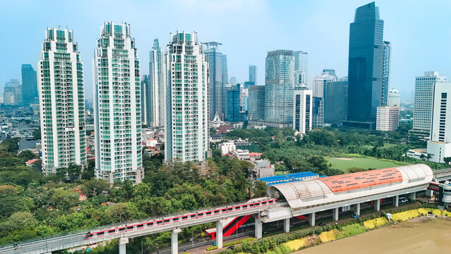 Aerial view of the modern Jakarta skyline featuring an elevated MRT train line passing through lush green areas and tall residential skyscrapers in Jakarta, Indonesia.