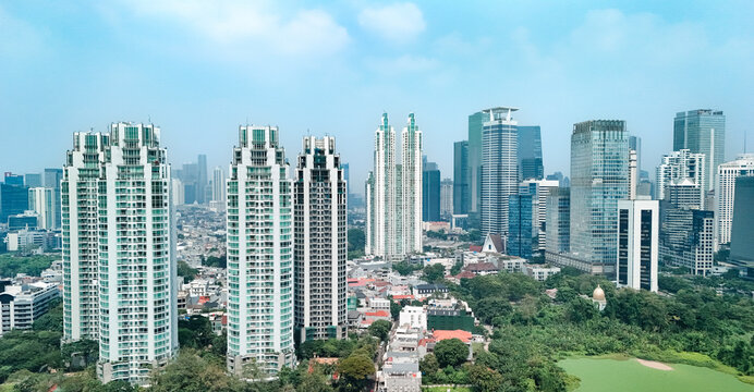 Aerial view of modern skyscrapers and high-rise buildings rising above lush green trees and urban parkland under a clear blue sky in the city center, Jakarta, Indonesia.
