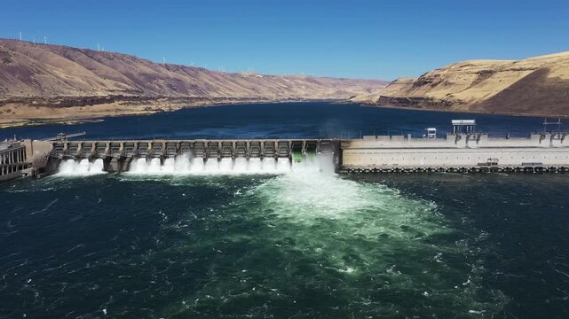 4K aerial drone footage of John Day Dam near Rufus, Oregon, USA with distant wind turbines. Hydroelectric power station, renewable energy, pelicans fishing in Columbia River Gorge