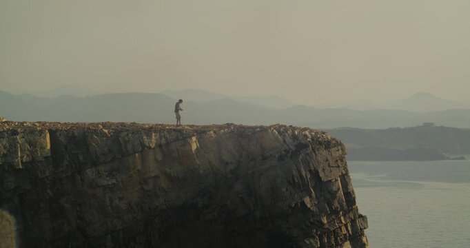 Dynamic wide shot of a lone man walking atop the dramatic cliffs of Cabo Pe&ntilde;as, Asturias, at golden hour, with rugged coastline, tranquil sea, and atmospheric misty mountains in soft sunlight.