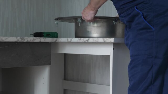 A professional installer carefully places a stainless steel round sink into the cutout of a marble countertop. Quality plumbing fixture installation during kitchen furniture assembly