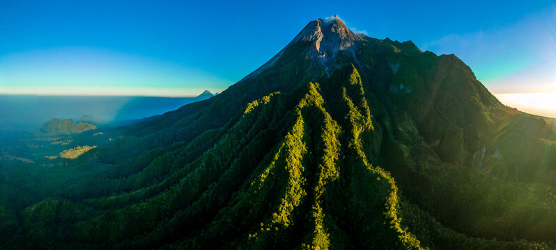 Aerial view of Mount Merapi with lush green volcanic slopes, rocky summit, and golden sunlight illuminating the landscape in Indonesia.