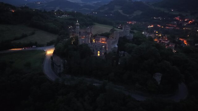 Aerial drone view of Rocchetta Mattei at twilight in Grizzana Morandi, Emilia-Romagna, Italy. Illuminated fortress with evening lights, golden hour glow, and cinematic 4K panorama over the Apennines.