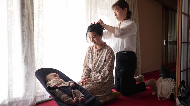 Soft light fills a quiet room. Woman tends to a baby's hair. Another woman sits nearby, smiling gently. Third woman brushes the first woman's hair. Both women wear traditional Japanese attire
