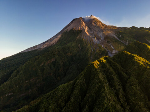 Aerial view of Mount Merapi with its volcanic peak emitting smoke above lush green forested slopes during golden hour in Indonesia.