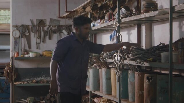 Young Indian Asian man or male fabricator or metal worker retrieving a bundle of coiled wire or vinyl cable from a storage rack containing raw material, tools, and raw material in fabrication workshop