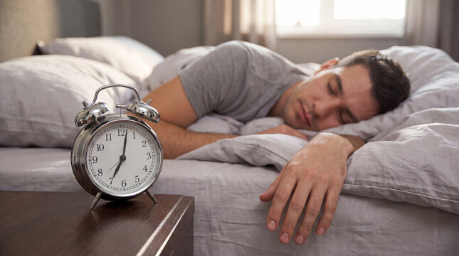Man sleeping in bed with gray sheets, alarm clock on nightstand showing time, soft natural light coming through window