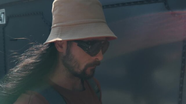 Authentic Bearded Man With Long Hair Wearing Bucket Hat And Sunglasses Walking Along Brick Wall On Sunny City Street Close Up Panning Portrait Of Casual Hipster Traveler During Summer Vacation