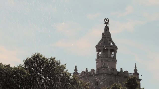 Heavy water drops falling in foreground of historic Stalinist empire style building tower with Soviet star on spire against blue sky and green trees in Eastern Europe during warm sunny summer day