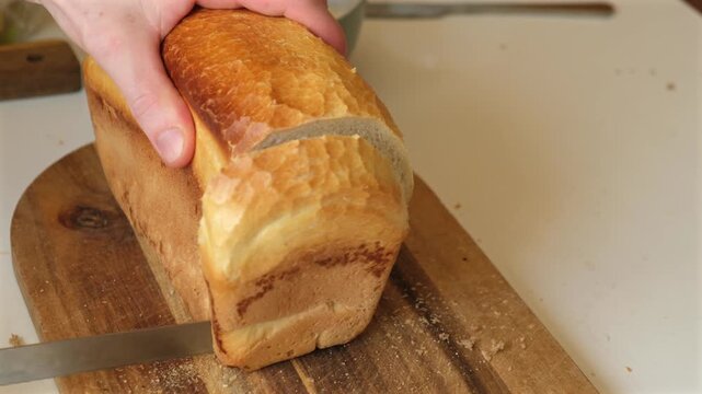 Close-up of a person slicing a fresh homemade white bread loaf with a bread knife on a rustic wooden cutting board. Fresh bakery, homemade baking, food preparation and breakfast concept.
