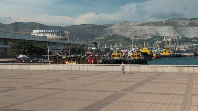 Unrecognizable small child walking alone on wide paved city embankment with massive negative space against sea port ships marine terminal and high quarry mountains in Novorossiysk on a summer day