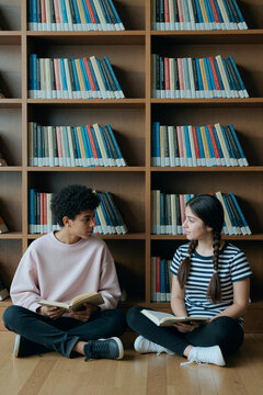 Vertical shot of multiethnic teenagers sitting on floor, reading books and discussing lesson in library. Education, literacy, study campaign, school marketing use