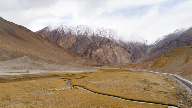 Aerial view of a winding stream flowing through an arid valley with snow-capped mountains in the background and a small group of people with livestock in Leh, Ladakh, India.