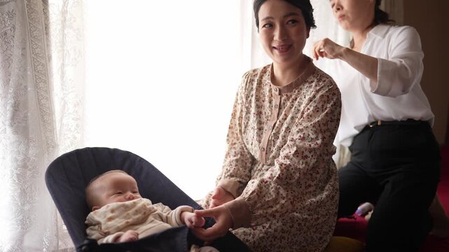 Soft light bathes a quiet family scene. Mother smiles gently while holding a sleeping infant. Third person leans close, perhaps adjusting or comforting. Warm, tender moment captured indoors