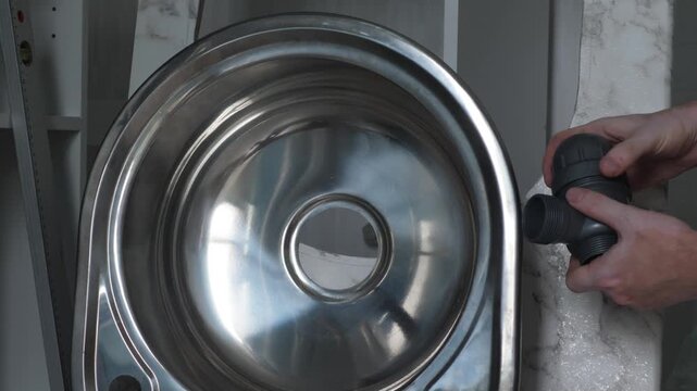 A plumber assembles a plastic siphon near a round stainless steel kitchen sink. Professional plumbing installation and drain pipe setup process during home renovation.