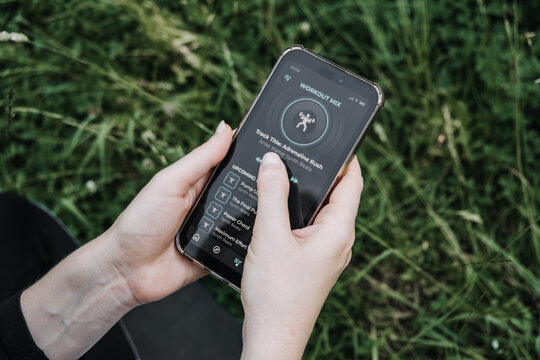Hands hold smartphone displaying workout mix music playlist application against green grassy background. Fitness motivation, digital lifestyle, audio streaming, active living.