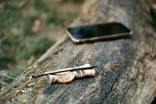Smartphone lies screen down on weathered wooden log next to incense stick. Phone left aside, digital detox nature, disconnecting from screen outdoor, unplugged nature moment.
