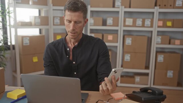 Man holding smartphone and checking laptop at desk amid stacked cardboard boxes on shelves in a building; small business focus.