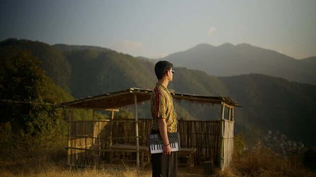 Alternate wide angle of a young man in patterned shirt holding a MIDI keyboard near a bamboo hut against a dramatic mountain skyline at dusk
