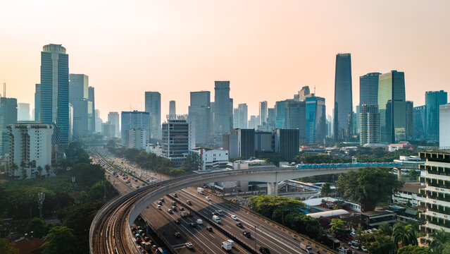 Aerial view of the modern Jakarta skyline featuring towering skyscrapers, a busy multi-lane highway with traffic, and elevated train tracks in Jakarta, Indonesia.