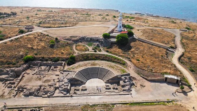 Aerial view of the ancient amphitheatre at the Archaeological Site of Nea Paphos in Cyprus. Stone seating, historic ruins, walking paths and the nearby lighthouse create a scenic coastal heritage view