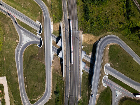 Aerial view of a highway interchange with curved roads and a train traveling on parallel railway tracks through a green landscape in Kaunas, Kaunas County, Lithuania.