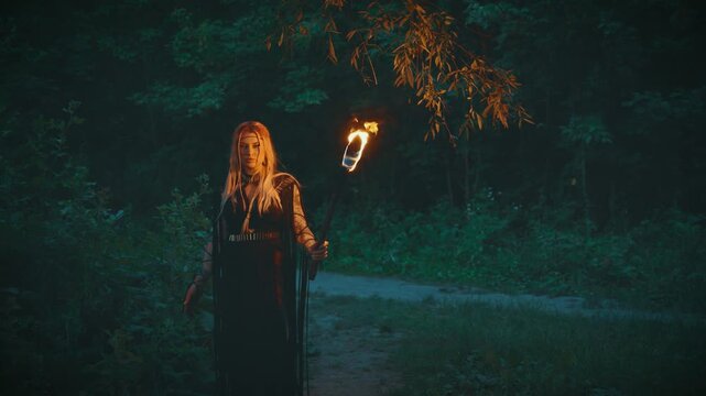 Gothic fantasy woman dark queen walking in mystical dusk forest trees holding burning torch in hands, bright fire flame lights way in darkness night. Sexy fairy Girl witch black long boho style dress.