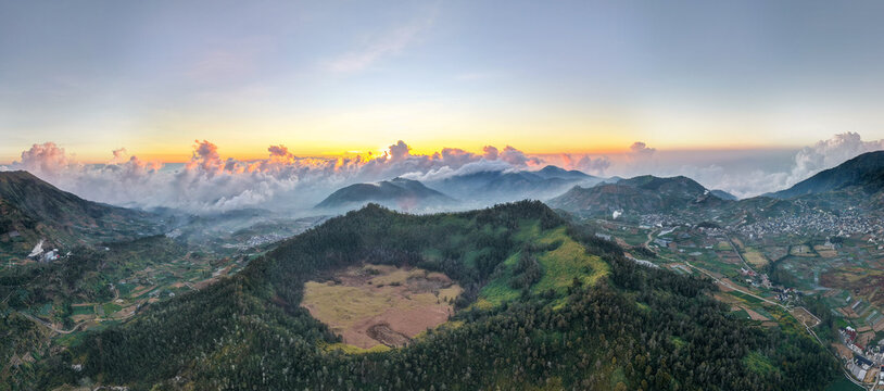 Aerial view of Dieng Plateau featuring a volcanic crater surrounded by lush green hills, agricultural terraces, and a dramatic sunrise sky in Indonesia.