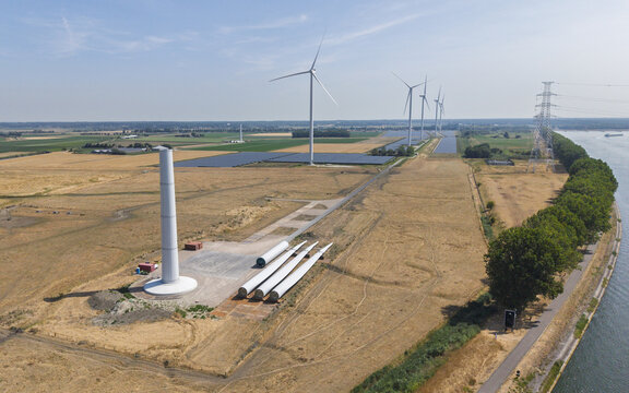 Aerial view of a wind farm construction site with turbine blades and a tower base on the ground, near operational turbines, solar panels, and a river in Ossendrecht, North Brabant, Netherlands.